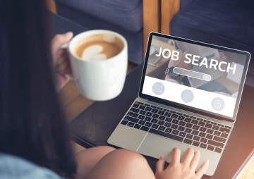 Žena drží v ruce kávu a vyhledává práci na notebooku